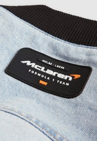 Kurtka jeansowa w jasnoniebieskim kolorze, z czarnym prążkowanym kołnierzem oraz teksturowaną czarną metką z napisem "McLaren Formula 1 Team" w kolorze białym i pomarańczowym.