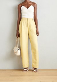 White ribbed tank top with chain straps, yellow high-waisted pants, beige handbag, and white block-heeled sandals on a wooden floor.