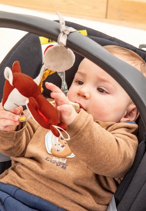Un jouet en peluche en forme de renard, marron et crème, solidement attaché à un siège auto pour bébé. La main du bébé atteint le jouet, montrant de l'intérêt.