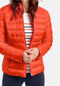 Röd puffjacka med quiltad struktur, frontdragkedja och sidofickor, buren över en vit skjorta med orange och svarta ränder.