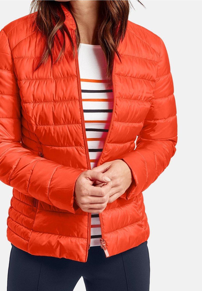 Röd puffjacka med quiltad struktur, frontdragkedja och sidofickor, buren över en vit skjorta med orange och svarta ränder.