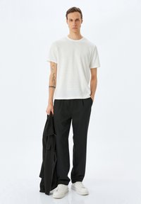 Vit, texturerad t-shirt med kort ärm, i kombination med svarta, lösa byxor och vita sneakers; håller en svart jacka och står framför en vit bakgrund.