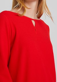 Chemisier rouge en tissu texturé, décolleté large avec un détail en trou de serrure et manches longues. La couleur est vive et unie.