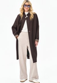 Kobieta w za dużym ciemnobrązowym płaszczu, beżowych szerokich spodniach, białej bluzce, białych trampkach i dużych kwadratowych okularach przeciwsłonecznych, stojąca na tle białego tła.