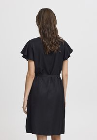 Schwarzes Kleid mit kurzen Rüschensäumen, tailliertem Gürtel und knielangem Rock. Glattes Gewebe mit subtiler Schattierung.