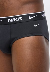 Κοντινό πλάνο ενός ατόμου που φοράει μαύρα εσώρουχα Nike με λευκό λογότυπο και ζώνη με σήμανση, σε απλό ανοιχτό φόντο.