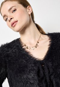 Jonge vrouw met een lichte huid en blond haar in een lage paardenstaart, gekleed in een zwarte fuzzy trui en een gouden charm-halsketting met verschillende bedels.
