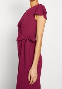 Vestido ajustado en un profundo magenta, con mangas de vuelo y un detalle anudado en la cintura. Tejido suave y liso con un diseño drapeado.