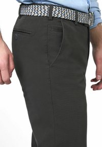 Mann in schwarzen Hosen, hellblauem, in die Hose gestecktem Hemd und einem gewebten Gürtel in Blau-, Beige- und Brauntönen, mit entspannten Händen an den Seiten.