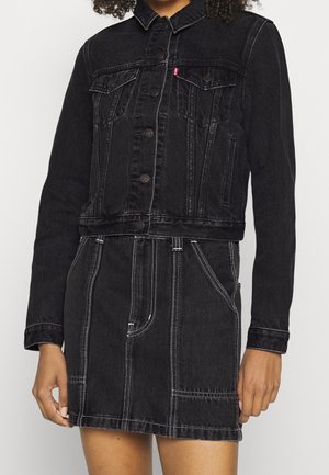 Kvinde iført en sort denimjakke med knapper over et matchende sort denimsærk med hvide syningsdetaljer.
