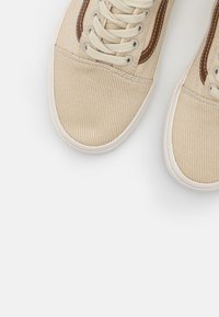 Baskets en toile beige avec semelles en caoutchouc blanches, lacets beiges et accents en cuir marron près des œillets sur fond blanc.