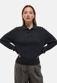 Pull en tricot gris foncé avec un col, des boutons et des poignets côtelés, associé à un pantalon à motif pied-de-poule.