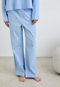 Ljusblå pyjamas med vertikala vita ränder. Tyget har en slät yta; byxorna är löst sittande med rak skärning och utan synlig hårdvara.