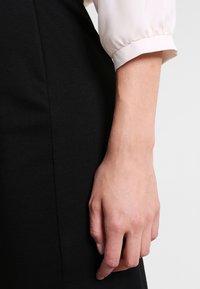 Svart kroppsnära klänning med ärmar i en kontrasterande ljus tyg, som har en slät textur, rena linjer och en skräddarsydd passform.