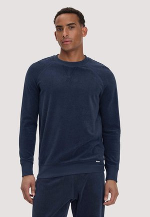 Mann trägt einen dunkelblauen Pullover mit Rundhalsausschnitt und langen Ärmeln sowie passende Hose, steht vor schlichtem hellem Hintergrund.
