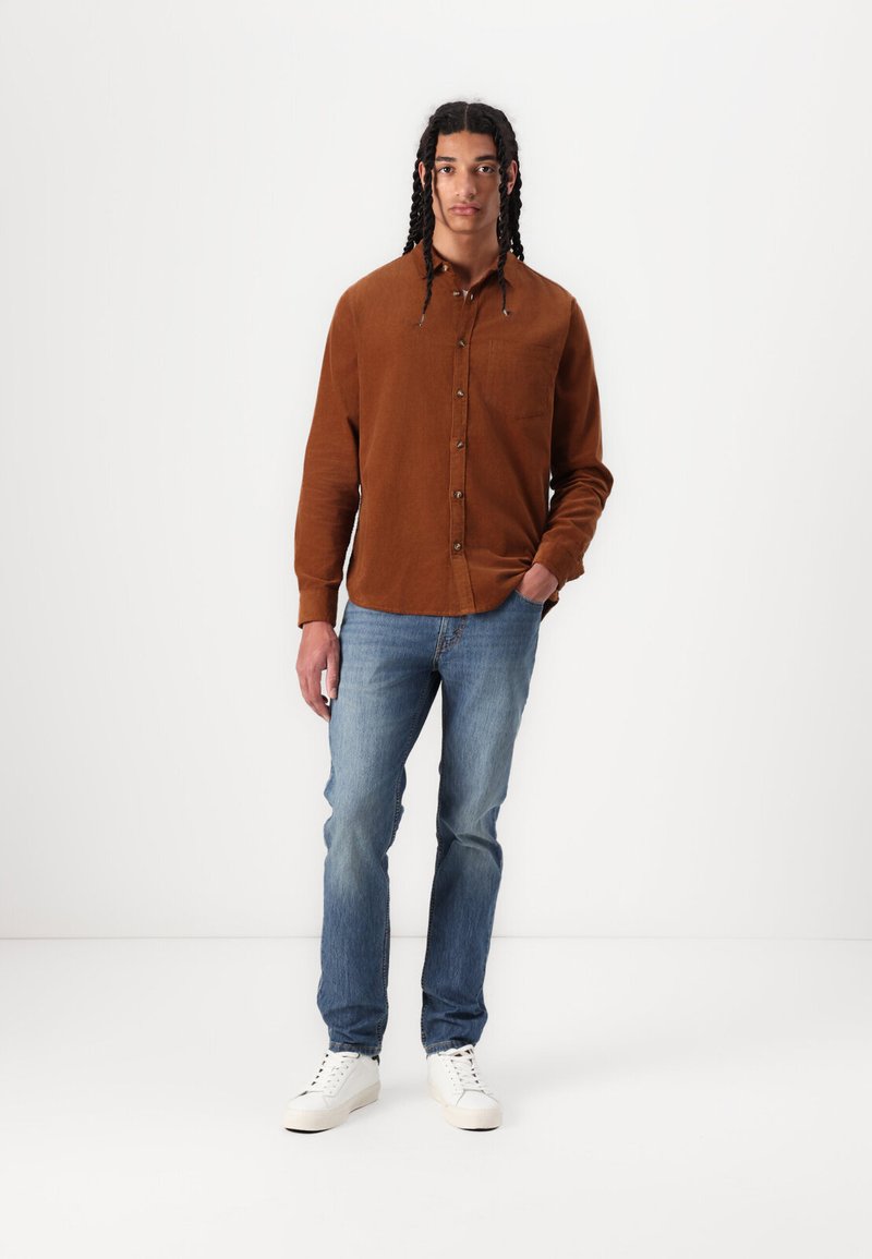Camisa marrón de botones con un bolsillo en el pecho, combinada con jeans azul claro y zapatillas blancas. El modelo está de pie frente a un fondo blanco liso.