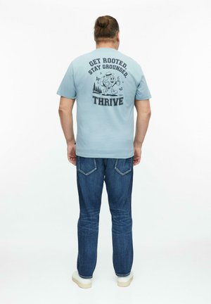 Mężczyzna z włosami związanymi w kok, ubrany w jasnoniebieski t-shirt z tekstem i grafiką "Get Rooted, Stay Grounded, Thrive" na plecach, niebieskie dżinsy i białe buty.