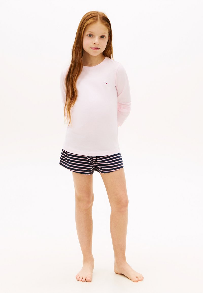 Jeune fille aux longs cheveux roux portant un t-shirt rose clair à manches longues et un short bleu marine rayé, debout pieds nus sur un fond blanc.