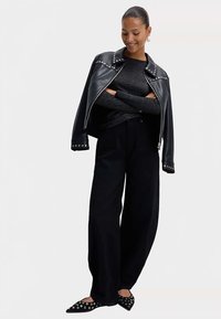 Schwarze Lederjacke mit silbernen Nieten, grauer strukturierter Pullover, schwarze weite Hose und schwarze spitze Ballerinas mit Ösenakzenten.