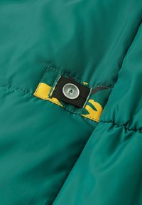 Détail d'une veste d'hiver verte avec un extérieur texturé, comportant un bouton et une patch en tissu avec un motif jaune et vert.