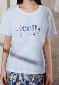 Ljusblå kortärmad t-shirt med en texturerad, flerfärgad "SeeBy Girl" grafik. Materialet verkar mjukt med en avslappnad passform.