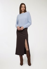 Donna che indossa un maglione blu chiaro in peluche, una lunga gonna marrone scuro con uno spacco sulla coscia, e stivali marroni fino al ginocchio, in piedi contro uno sfondo bianco.