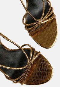 Metaliczne złote sandały na wysokim obcasie z teksturą wężowej skóry, z skrzyżowanymi paskami i zaprojektowane w szpic przy czarnej podeszwie.