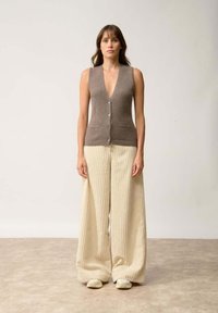Brązowy, bezrękawnik z głębokim dekoltem w kształcie V i guzikami z przodu, w połączeniu z szerokimi, kremowymi spodniami o prążkowanej strukturze i jasnym obuwiem.