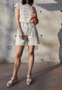 Camisa blanca de manga corta con rayas rojas, pantalones cortos de mezclilla beige y sandalias gruesas beige. Sosteniendo un pequeño bolso naranja. Fondo de hormigón.