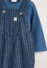 Niebieska prążkowana koszulka z długim rękawem pod ciemnoniebieskimi sztruksowymi ogrodniczkami w kratę z przednią kieszenią i drewnianymi guziczkami na szelkach dla niemowlaka.