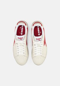 White sneakers with a textured finish, featuring red accents and logo, round laces, and a smooth rubber sole. Classic athletic design.