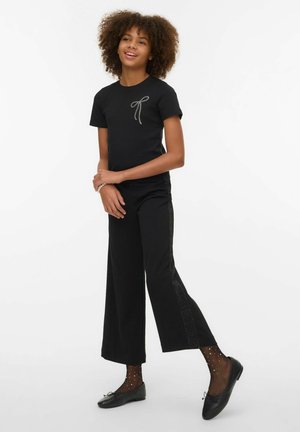 Black short-sleeve t-shirt with a bow design, paired with wide-leg black trousers featuring glittery side accents. Black shoes and patterned socks.