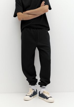 Personne portant un pantalon de survêtement noir, un t-shirt noir, des chaussettes blanches avec des rayures noires et des baskets noires et blanches, debout les bras croisés.