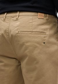 Gros plan sur la poche arrière d'un pantalon beige, ornée de trois petites croix brodées et d'une étiquette en cuir indiquant "Scotch & Soda Amsterdam".