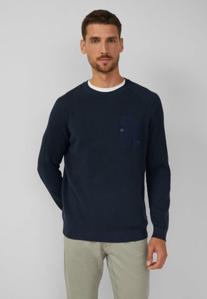 Marineblaues Strickpullover mit strukturiertem Muster, Rundhalsausschnitt und einer aufgesetzten Tasche auf der linken Brust. Kombiniert mit hellen Hosen.