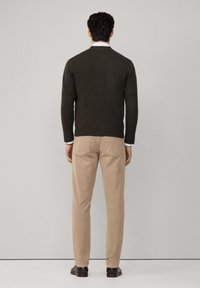 Muškarac okrenut leđima, odjeven u tamnozeleni pulover, s vidljivim bijelim rukavima košulje, bež hlačama i tamnosmešnim cipelama, s jednostavnom pozadinom.