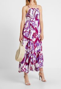 Maxi šaty s fialovým a růžovým květinovým vzorem, smocková sukně, tenké ramínka, nabíraná sukně, v kombinaci s tkanou taškou a stříbrnými vysokými podpatky.