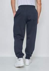 Pantalons de survêtement navy avec une taille élastique et des poignets élastiques, dotés d'une poche arrière. Portés avec des baskets blanches et des chaussettes blanches visibles.
