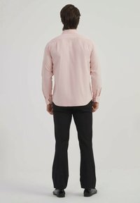 Chemise rose clair à manches longues, avec un empiècement au dos et un ourlet arrondi, associée à un pantalon noir et des chaussures. Toucher de tissu lisse.