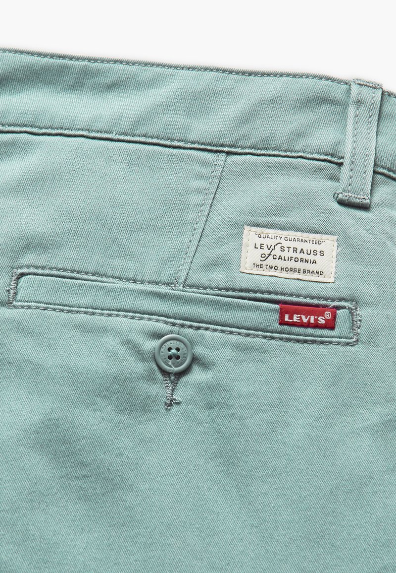 Pantalones de mezclilla azul claro con un bolsillo trasero, que cuenta con un botón gris y dos etiquetas: una blanca con texto negro y la otra roja con texto blanco.