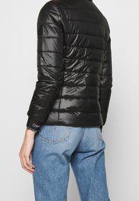 Sort puffer-jakke med quiltet design, skinnende tekstur og en rund skulderpatch. Manchetter har et hvidt logoakcent på en mørk baggrund.