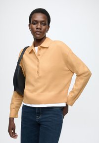 Gaiši oranžs adīts džemperis ar apkakli, pogām priekšpusē un ribotu apakšu. Kopā ar tumši zilām džinsām un melnu plecu somu.