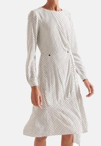 Biała sukienka z czarnym abstrakcyjnym wzorem, długie rękawy, okrągły dekolt, dopasowana talia i drapowana spódnica z asymetrycznym obszyciem.