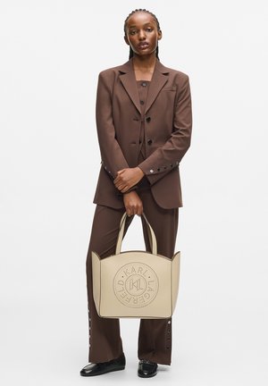 Woman in brown suit holding beige tote bag with circular perforated logo, standing against plain white background.