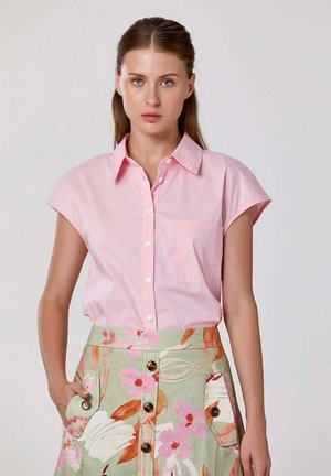 Femme portant une chemise rose clair à manches courtes boutonnée et une jupe taille haute à imprimé floral vert, rose et orange avec des poches avant.