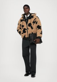 Beige sherpa jack met zwart abstract patroon, voorrits en capuchon. Gecombineerd met zwarte broek en een zwarte tas vastgehouden.