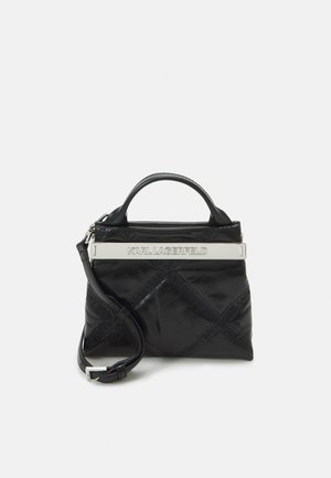 Musta pehmeä nahkalaukku lyhyellä kahvalla ja irrotettavalla hihnalla. Ylhäällä on hopeinen logo-levy. Sujuva pinta.