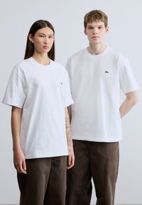 Λευκά βαμβακερά t-shirts με πράσινο λογότυπο Lacoste στο στήθος, σε συνδυασμό με καφέ παντελόνια. Χαλαρή εφαρμογή, κοντά μανίκια και σχέδιο με στρογγυλή λαιμόκοψη.