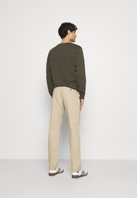Sweatshirt verde închis, pantaloni bej și adidași negri cu alb. Ținuta are o croială lejeră, cu cusături vizibile și detalii cu buton.