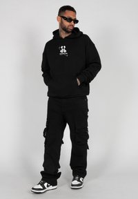 Svart hettegenser med lommer foran og hvit grafisk design, kombinert med svarte cargo-bukser med flere lommer. Joggesko med hvite detaljer.